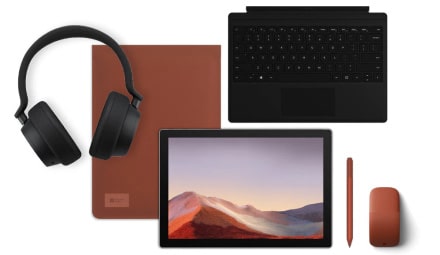 A flat lay of tech items including black headphones, a brown notebook, a tablet displaying a landscape, a black keyboard, a brown stylus, and a brown wireless mouse on a white background.
